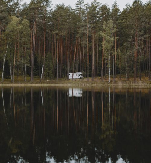 Camper van amidst a tranquil forest by a lake reflecting trees and sky in Lithuania.